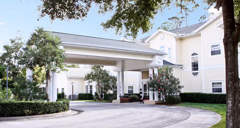 Front exterior of a light-colored senior living building with a covered porte-cochere, circular driveway, and landscaped trees and shrubs.