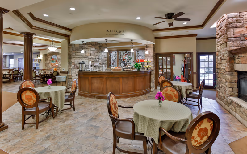 A cozy common area in a senior living facility with round tables covered in light green tablecloths, each adorned with a small vase of purple flowers. The room features a wooden reception desk with a stone backdrop and a 'WELCOME' sign above it. There are wooden chairs with floral upholstery around the tables, a stone fireplace on the right, and ceiling fans overhead. The space is warmly lit and inviting.