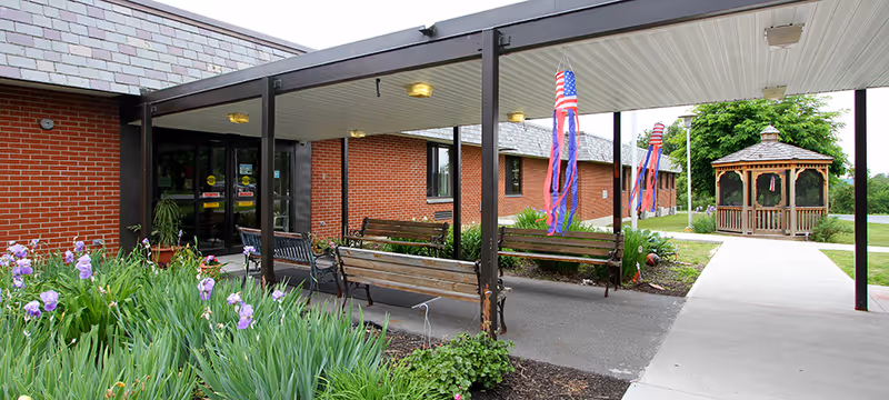 Entrance to a brick building with a covered walkway supported by metal posts. There are wooden benches along the walkway and colorful windsocks hanging from the posts. To the right, a paved path leads to a wooden gazebo surrounded by green grass and trees. Purple flowers and green plants are visible in the garden bed near the entrance.
