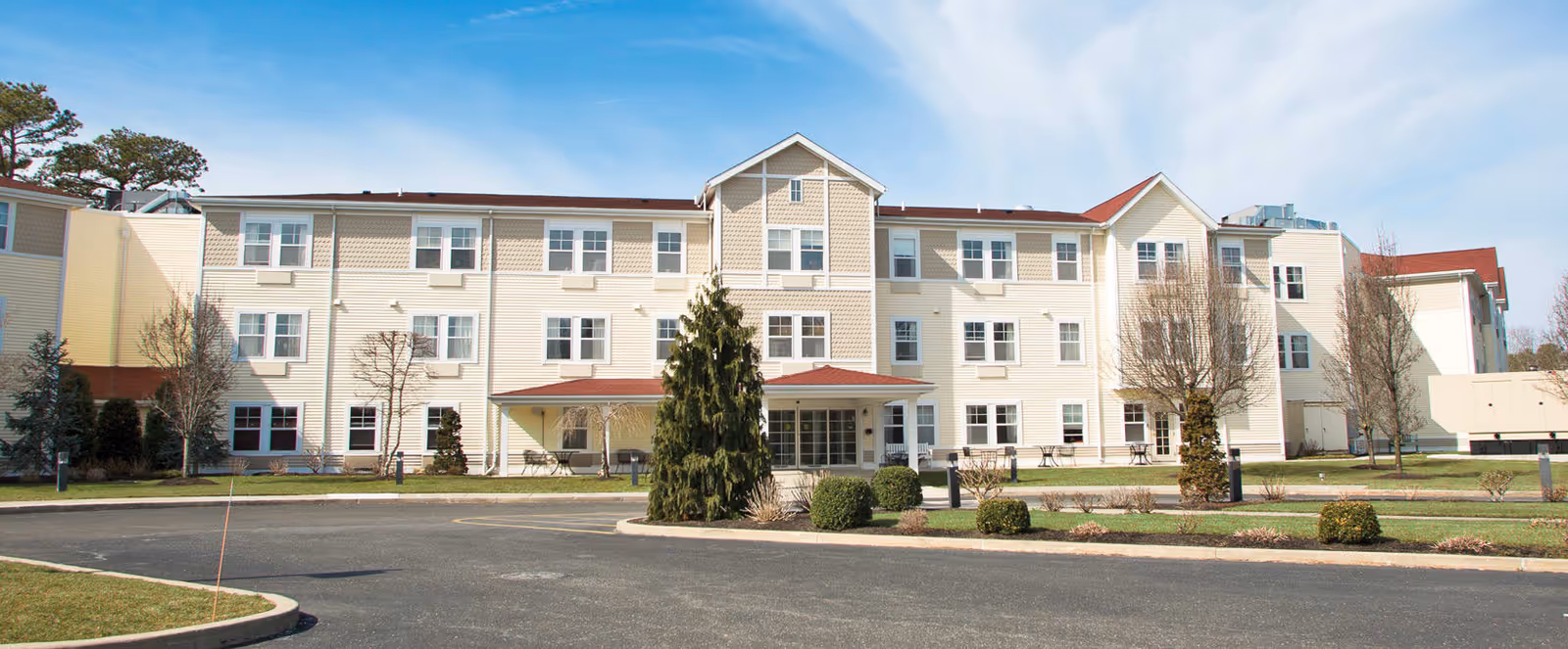 Exterior view of a three-story senior living facility building with beige siding, multiple windows, and a central entrance with a small covered porch. The building is surrounded by a paved driveway, landscaped bushes, and trees under a clear blue sky.