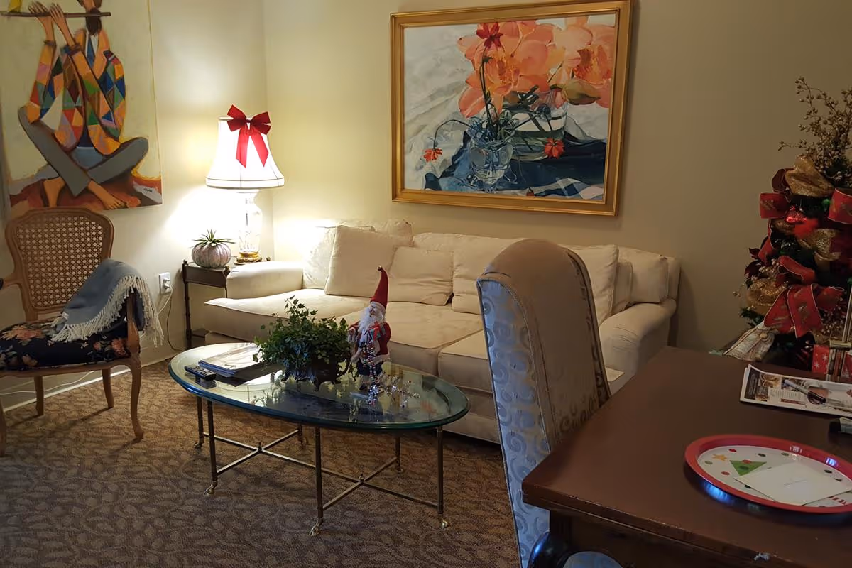 A cozy living room with a beige sofa, a glass coffee table with a small plant and a Santa Claus decoration, a wooden chair with a floral cushion and a blue throw, a table with a red tray and some papers, a lamp with a red bow, and two colorful paintings on the walls.