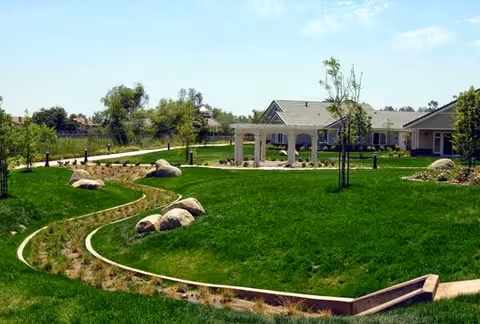 Well‑maintained green lawn with rock accents, a winding planted channel, walkways, and a small pavilion in front of single‑story residential buildings.