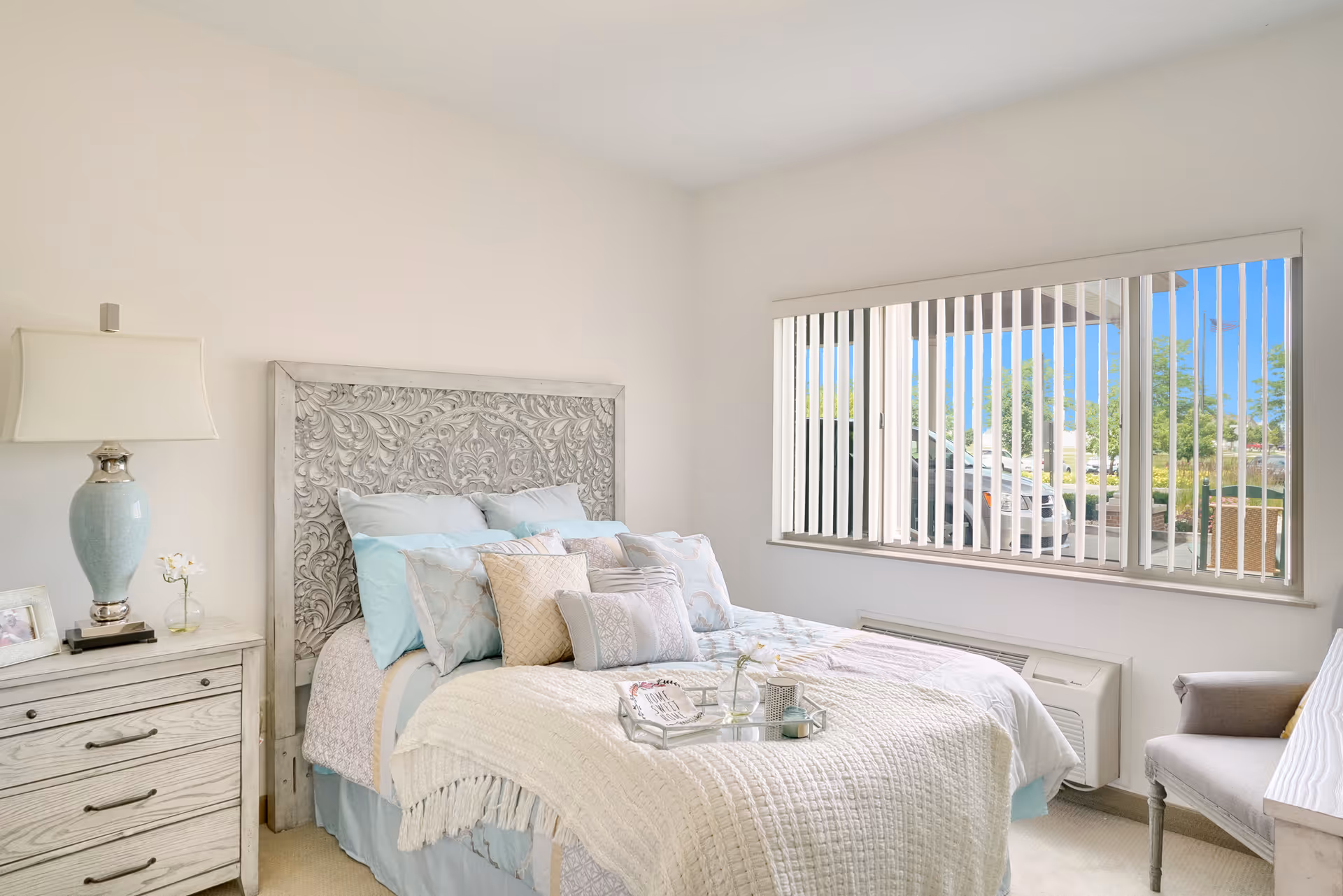 A bright bedroom with a decorative carved wooden headboard, a bed made with multiple pillows and a white textured blanket. There is a nightstand with a blue lamp and a small flower vase, a window with vertical blinds showing a view of parked cars outside, and a gray upholstered chair near the window.