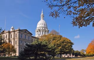 A large historic government building with a prominent dome, surrounded by trees with autumn foliage under a clear blue sky.