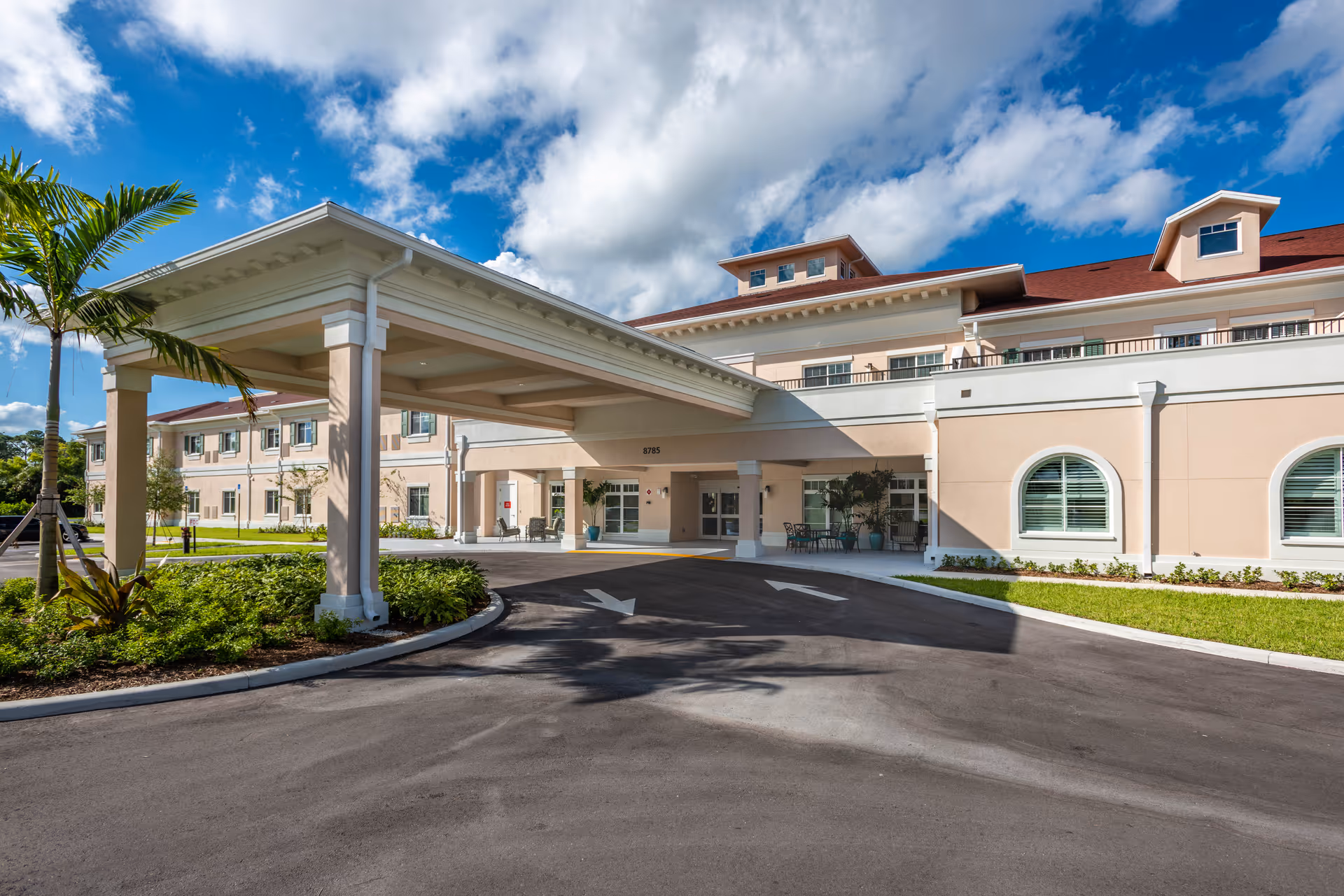 Exterior front of a senior living building with a covered entrance, driveway, and palm tree under a blue sky.