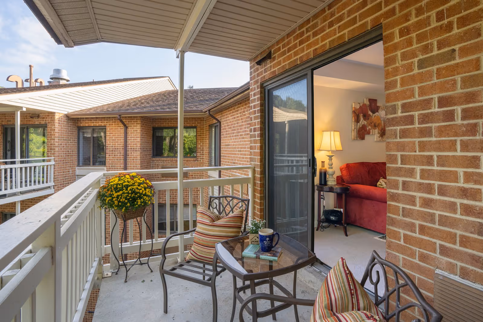 A small balcony with a glass-top table and two metal chairs with striped cushions. A potted plant with yellow flowers is placed on a stand near the railing. The balcony overlooks a brick building with windows. A sliding glass door leads inside to a room with a red couch, a side table with a lamp, and a floral painting on the wall.