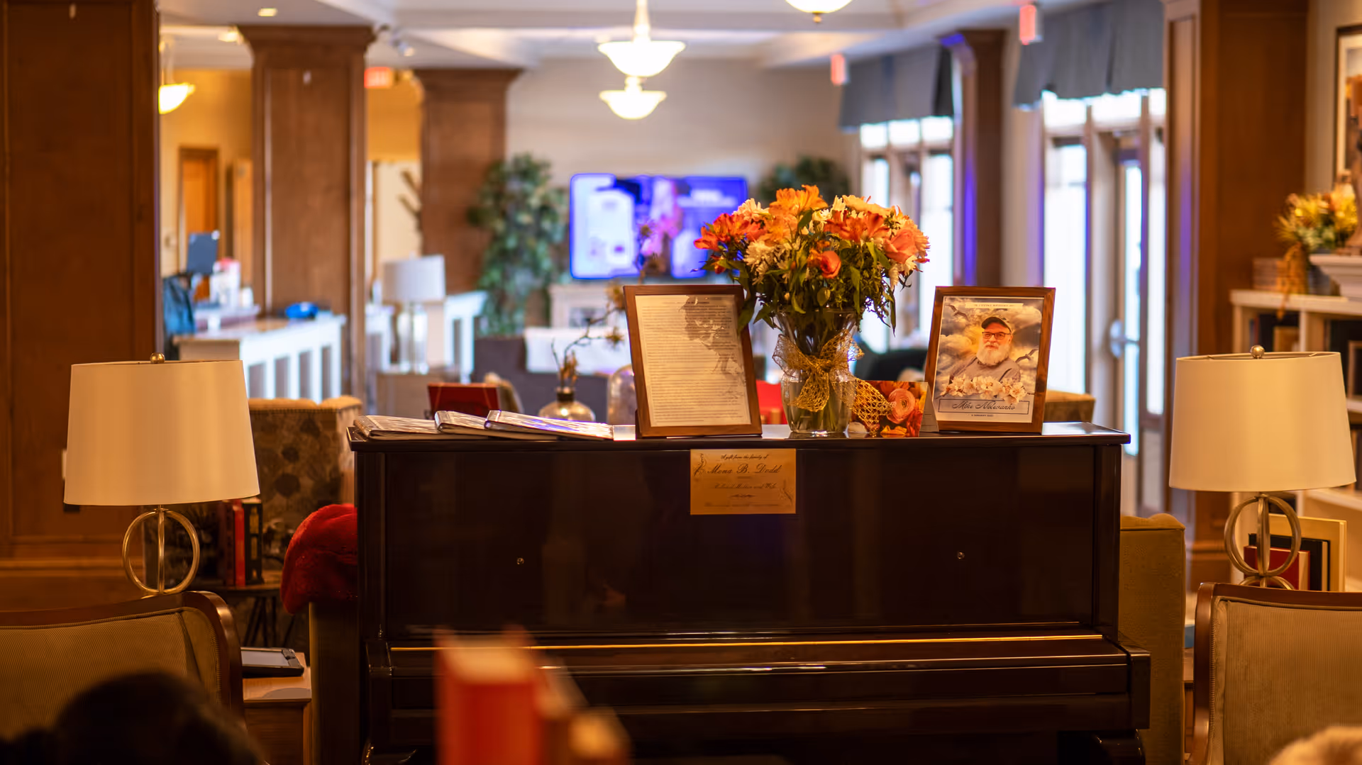 Cozy senior living lounge featuring a piano topped with flowers and framed photos, flanked by lamps and seating.