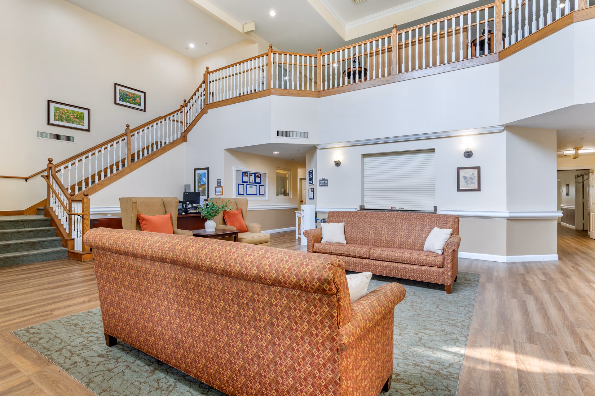 A spacious senior living facility common area with two patterned sofas and two beige armchairs arranged around a small wooden table with a plant. The room features wood flooring, a green patterned rug, and a staircase with wooden handrails and white balusters leading to an upper level. The walls are decorated with framed artwork and there is a reception window with a closed shutter.