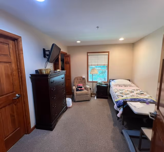 A small, cozy bedroom with a single bed covered with a colorful quilt, a beige armchair, a dark wooden dresser with a wall-mounted TV above it, a nightstand with a lamp, and a window with blinds letting in natural light.