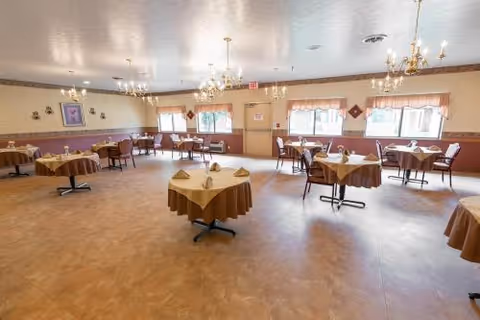 A spacious dining room with multiple round tables covered with brown tablecloths, each set with folded napkins. The room has large windows with valance curtains, chandeliers hanging from the ceiling, and a light-colored floor.
