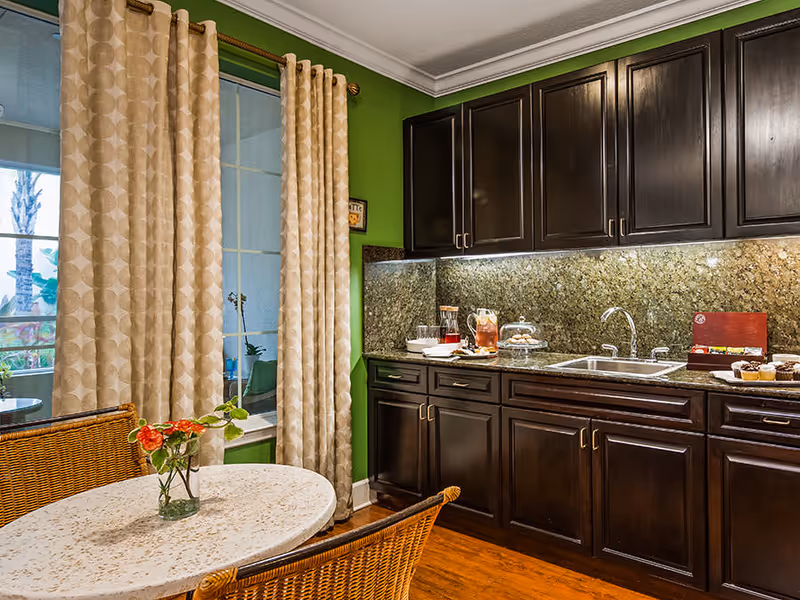 A cozy kitchen area with dark wooden cabinets, a granite countertop and backsplash, a stainless steel sink, and a small round table with a vase of flowers. Beige patterned curtains cover a window, and wicker chairs are placed around the table.
