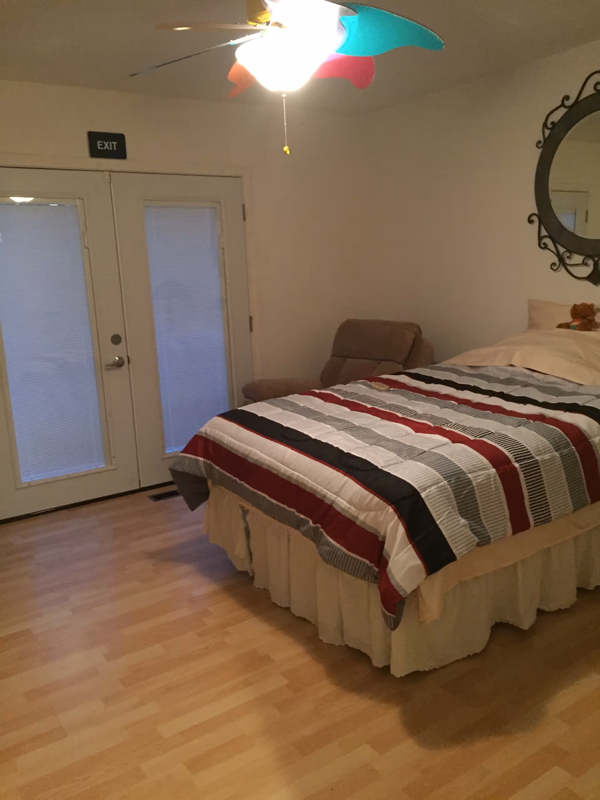 A bedroom with a bed covered in a striped comforter in shades of red, black, gray, and white. There is a beige armchair next to the bed and a round decorative mirror on the wall above the bed. Double doors with blinds and an exit sign above them are visible on the left side. A ceiling fan with colorful blades is on the ceiling.