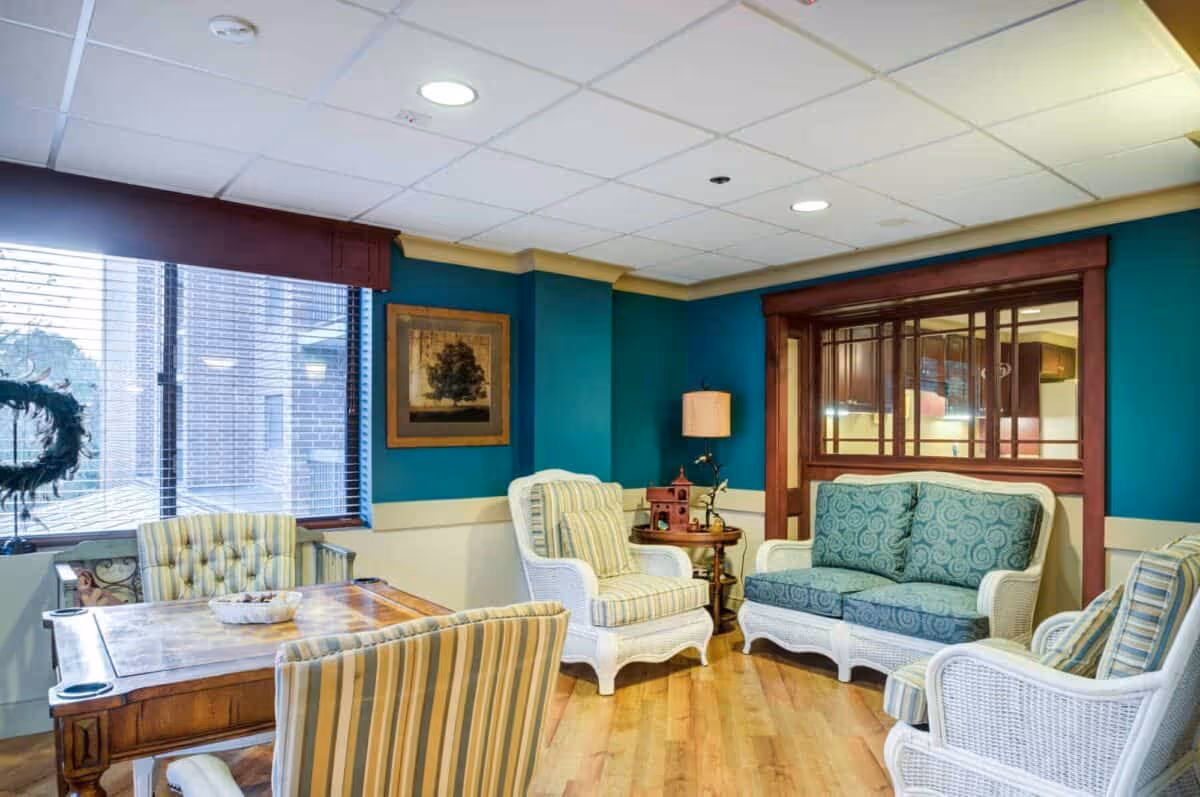A cozy sitting area in a senior living facility with a wooden table surrounded by striped upholstered chairs, white wicker armchairs with cushions, and a matching loveseat. The walls are painted teal with beige trim, and there is a framed picture on the wall. A window with blinds lets in natural light, and a lamp sits on a small round table in the corner.