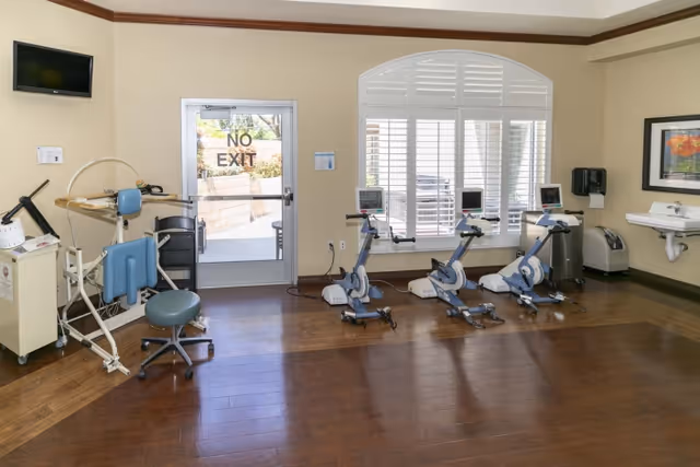 A rehabilitation or exercise room with three stationary exercise bikes lined up in front of a large window with white shutters. To the left, there is exercise equipment including a chair and a machine, and to the right, a wall-mounted sink and a hand sanitizer dispenser. A door with a 'NO EXIT' sign is visible in the background.
