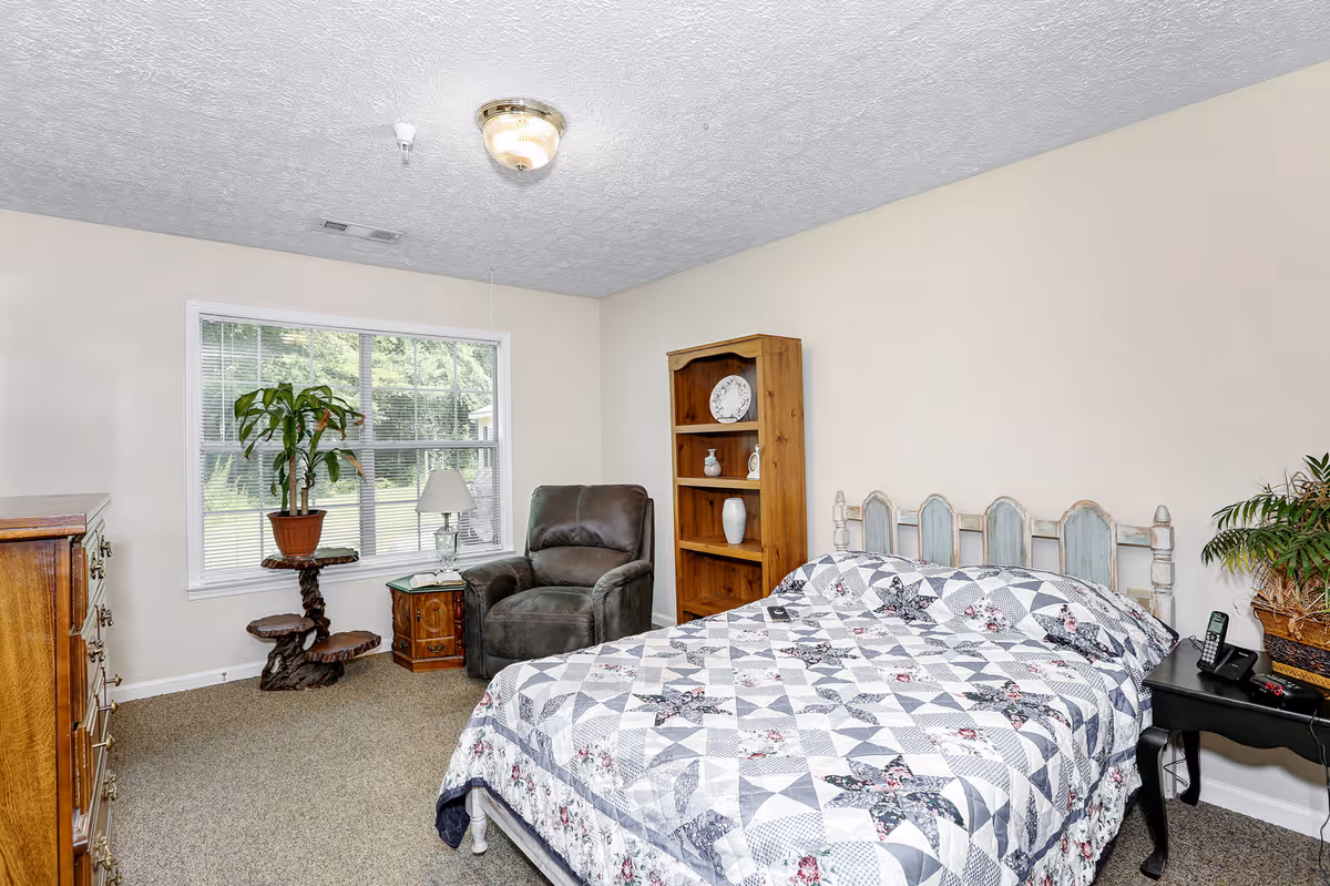 A bedroom with a bed covered in a patterned quilt, a wooden bookshelf with decorative items, a dark armchair, a small side table with a lamp, a wooden dresser, a plant on a unique wooden stand, and a large window showing greenery outside.