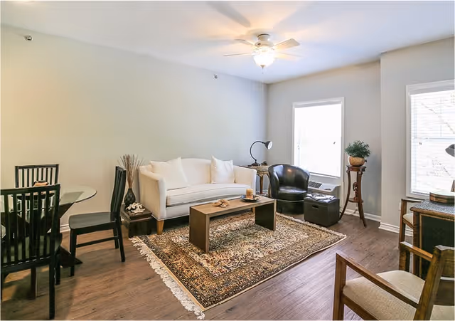 A bright living room with a white sofa, a black leather chair, a wooden coffee table on a patterned rug, a round glass dining table with black chairs, and two windows letting in natural light.