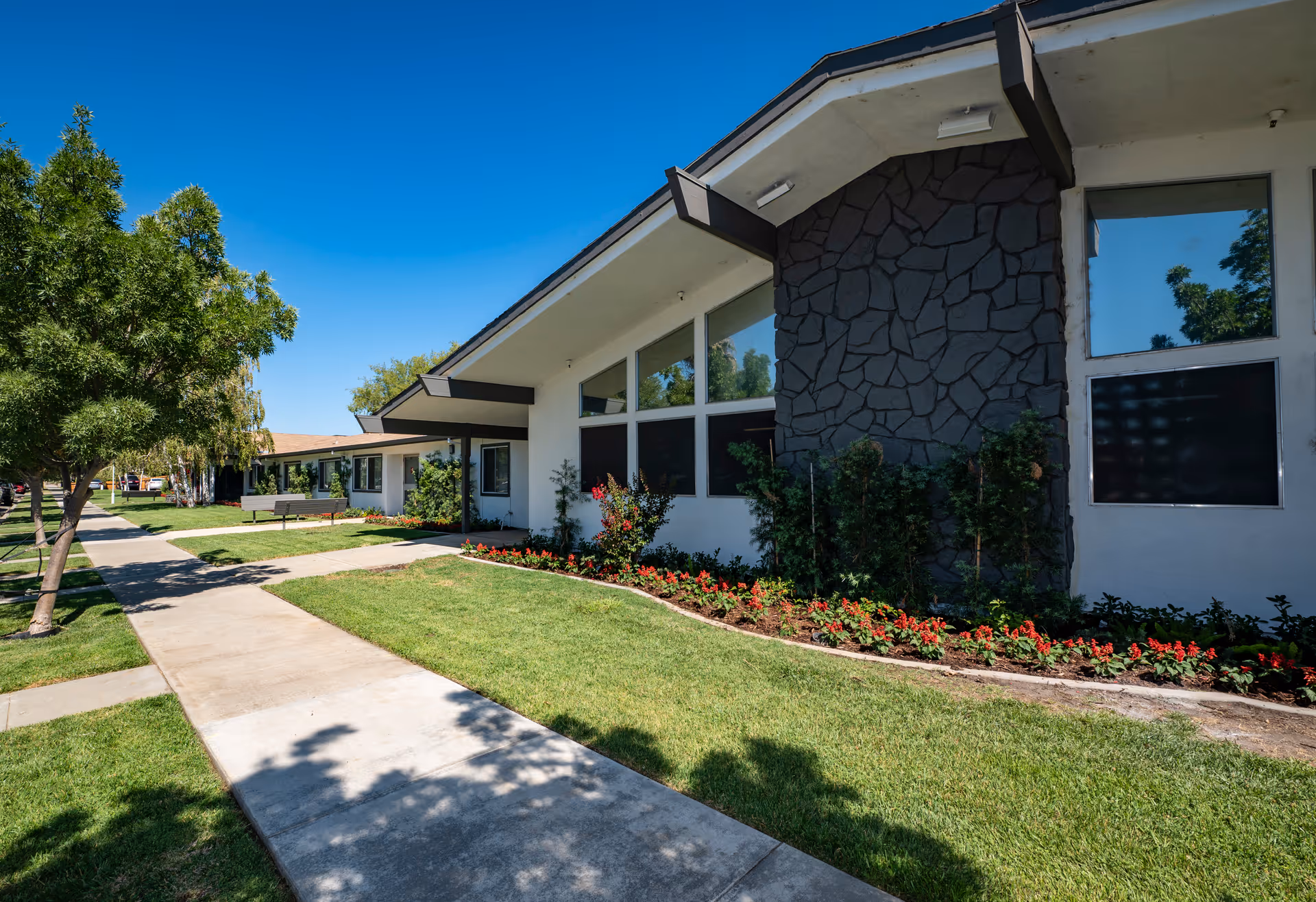 Exterior view of a single-story building with white walls and a stone accent wall, surrounded by well-maintained green grass, flower beds with red flowers, and trees along a concrete sidewalk under a clear blue sky.