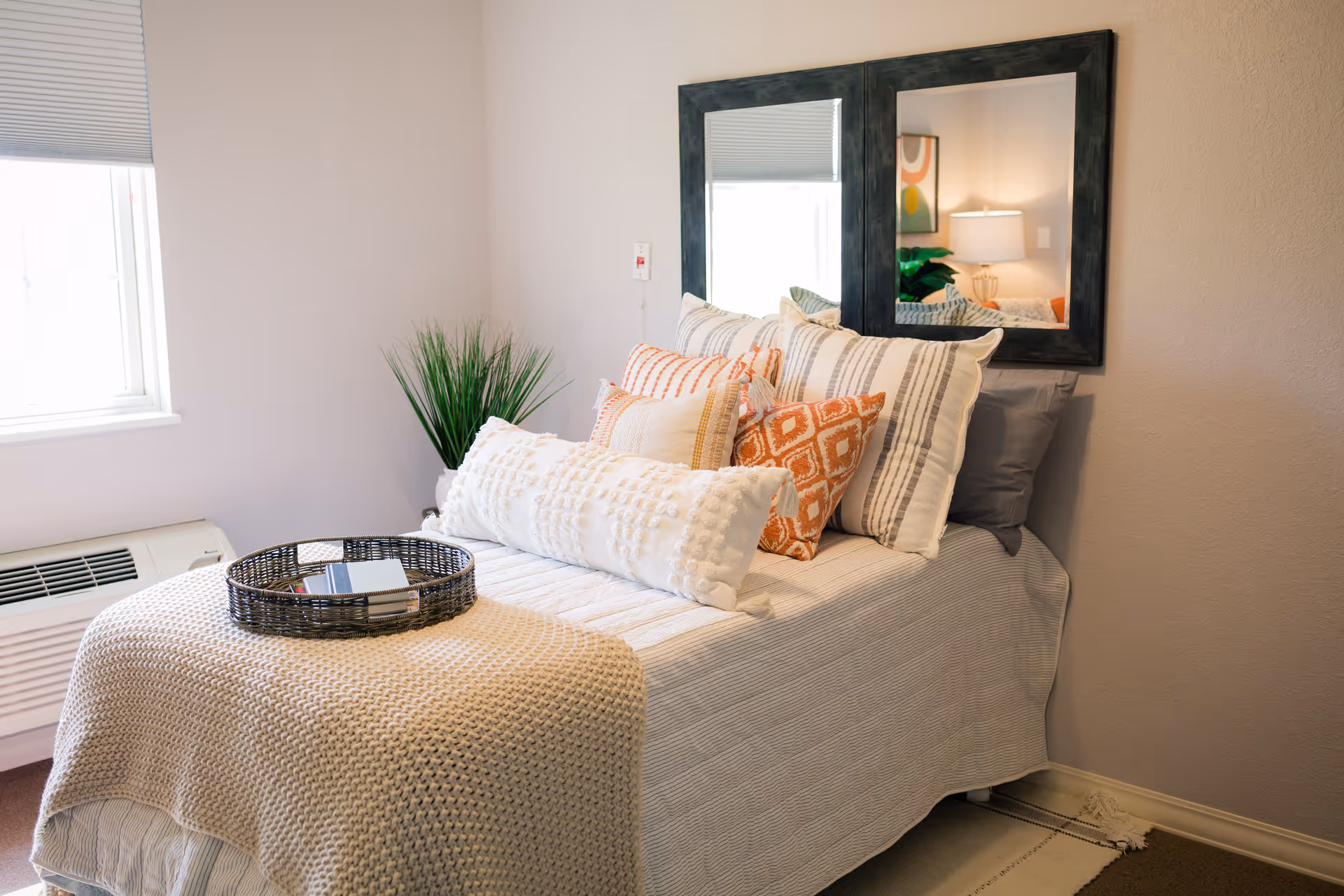 A neatly made bed with multiple decorative pillows in white, orange, and beige tones, a beige knitted throw blanket, and a round wicker tray with a book on top. The bed is positioned against a wall with two black-framed mirrors above it. A green plant is placed in the corner near a window with closed blinds, and a heating/cooling unit is below the window.