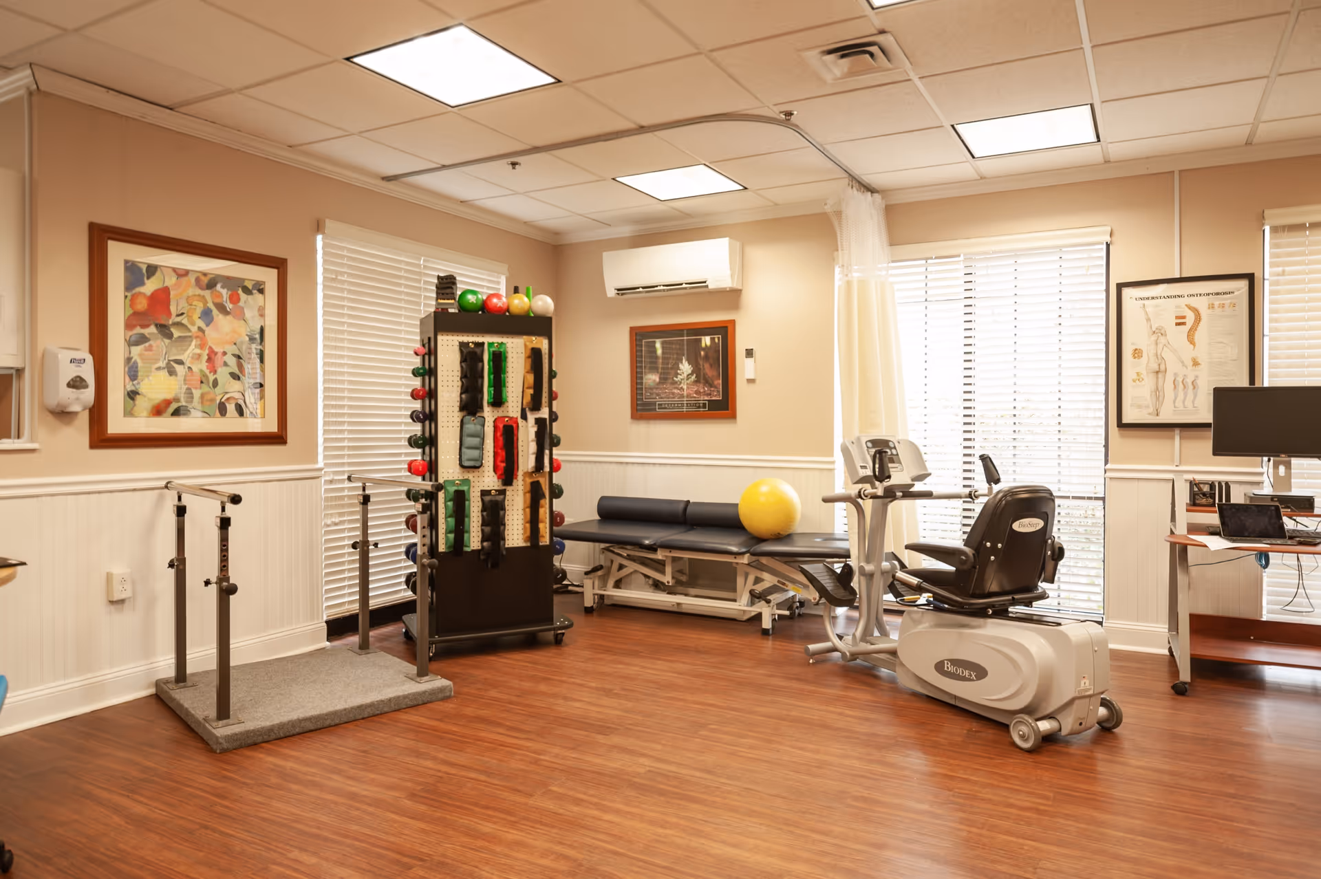 A well-lit physical therapy or exercise room with wooden flooring, featuring a recumbent exercise bike, parallel bars for walking support, a padded therapy table with a yellow exercise ball on it, and a rack holding various exercise equipment such as weights and resistance bands. The room has beige walls with white wainscoting, framed artwork, large windows with blinds, and a computer workstation in the corner.