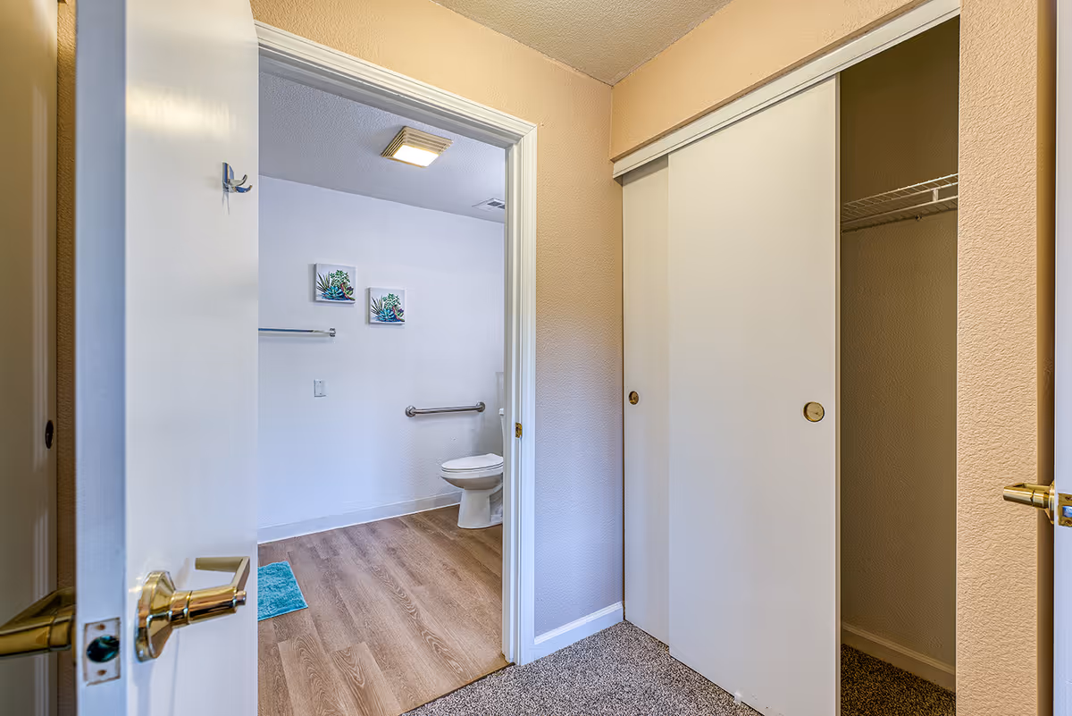 View from a carpeted room into a bathroom with wood-look flooring. The bathroom has a white toilet, a grab bar on the wall, two small framed pictures with plant designs, and a towel rack. To the right of the doorway is a closet with sliding white doors, one door partially open showing a wire shelf inside.