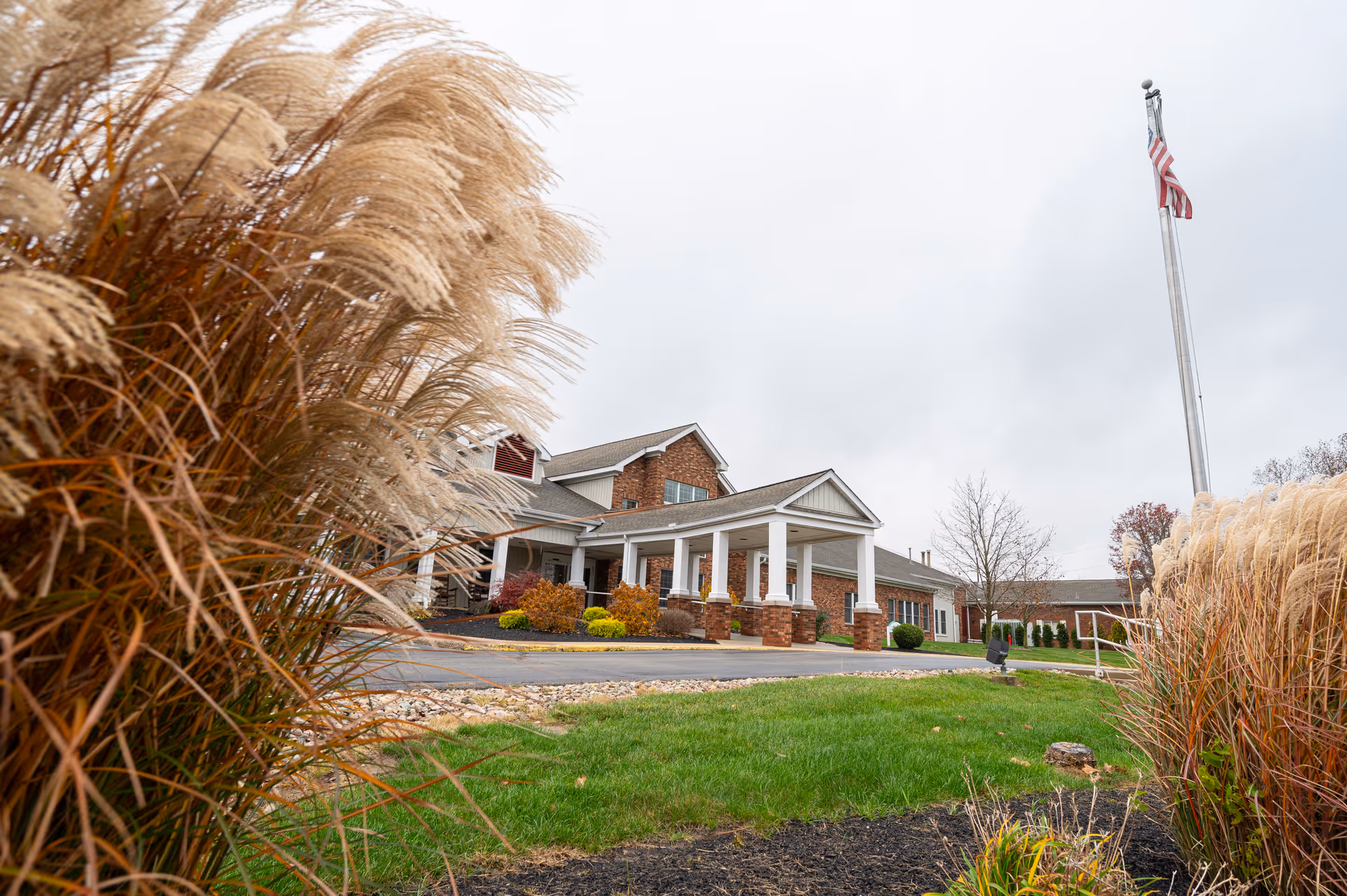 Front entrance of a brick senior living building with white columns, landscaped grounds and an American flag, seen through ornamental grasses.