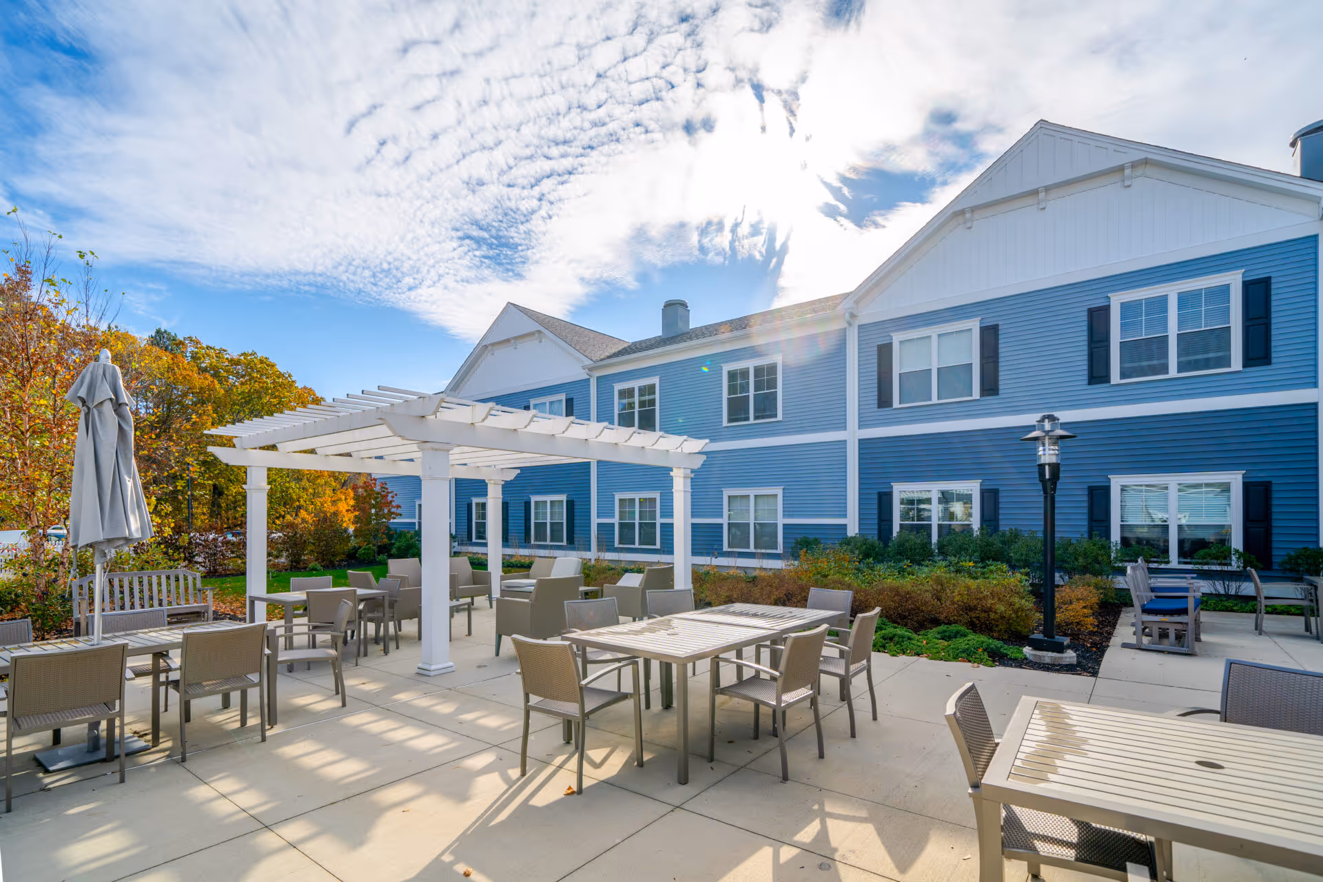 Outdoor patio area at The Branches of Framingham with multiple tables and chairs arranged under a white pergola and around the concrete patio. The building exterior is blue with white trim and black shutters. Trees with autumn foliage are visible in the background under a partly cloudy sky.