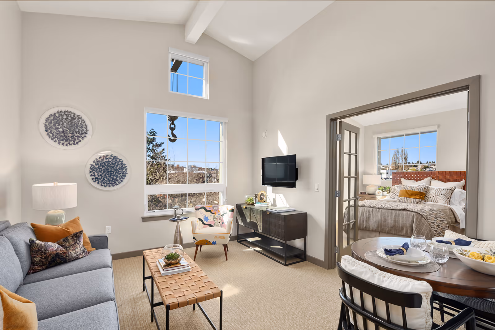 Sunlit living room with a gray sofa, coffee table, wall-mounted TV and dining table, with a bedroom visible through glass-paneled doors.