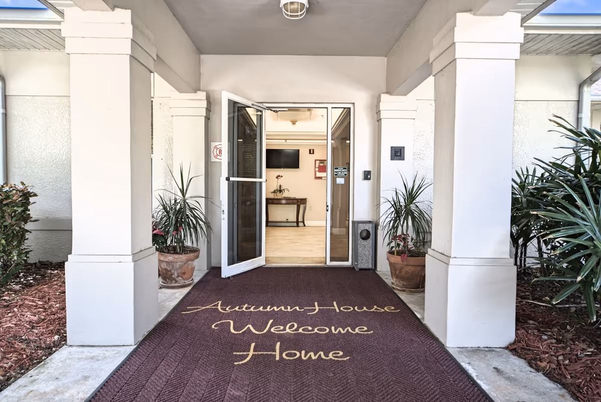 Entrance to Autumn House facility showing an open glass door with a maroon welcome mat that reads 'Autumn House Welcome Home'. The entrance is flanked by white pillars and potted plants on either side.