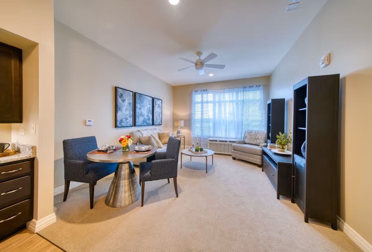 A bright and cozy living room area with beige walls and carpet. The room features a round dining table with four dark upholstered chairs, set with plates and a vase of flowers. In the background, there is a beige sofa with cushions, a coffee table with decorative items, a cushioned armchair near a large window with sheer curtains, and two dark wooden shelving units. A ceiling fan with lights is mounted on the ceiling.