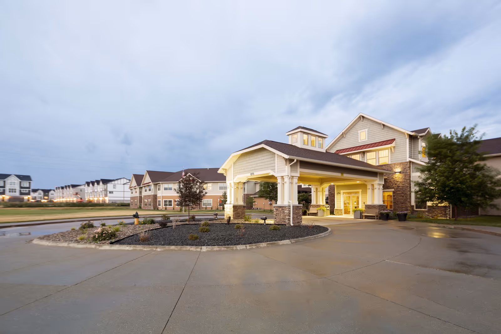Entrance canopy and driveway of a multi-story senior living facility with a landscaped roundabout under a cloudy sky.