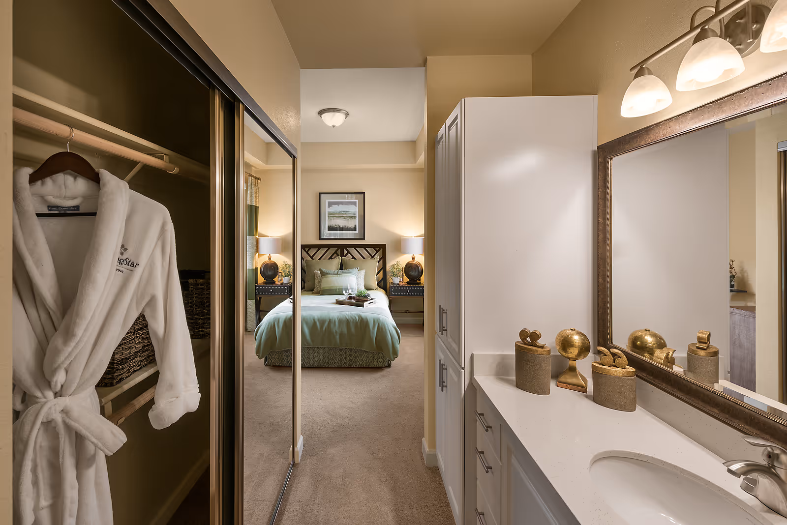 View of a bedroom from an adjacent bathroom area. The bedroom has a neatly made bed with green bedding, two bedside tables with lamps, and a framed picture on the wall. In the foreground, there is a bathroom vanity with a sink, countertop, decorative items, and a large mirror. To the left, a closet with sliding mirrored doors is partially open, showing a white robe hanging inside.