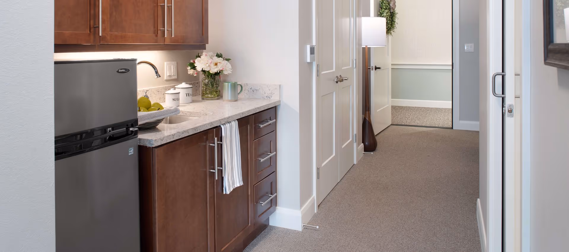 A small kitchenette area with dark wood cabinets, a marble countertop, a stainless steel mini refrigerator, a sink, and a vase of flowers. There are containers labeled sugar and tea, a striped towel hanging from a cabinet handle, and a bowl of green apples on the counter. The kitchenette opens into a carpeted hallway with white doors and a tall floor lamp with a wreath hanging above it.