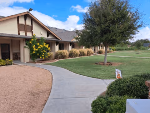 Exterior view of a single-story senior living facility building with a sloped roof, surrounded by a landscaped yard with green grass, bushes, a tree, and a curved concrete walkway leading to the entrance. The sky is partly cloudy.