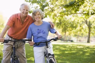 An elderly couple smiling and standing outdoors with their bicycles in a green park with trees in the background.