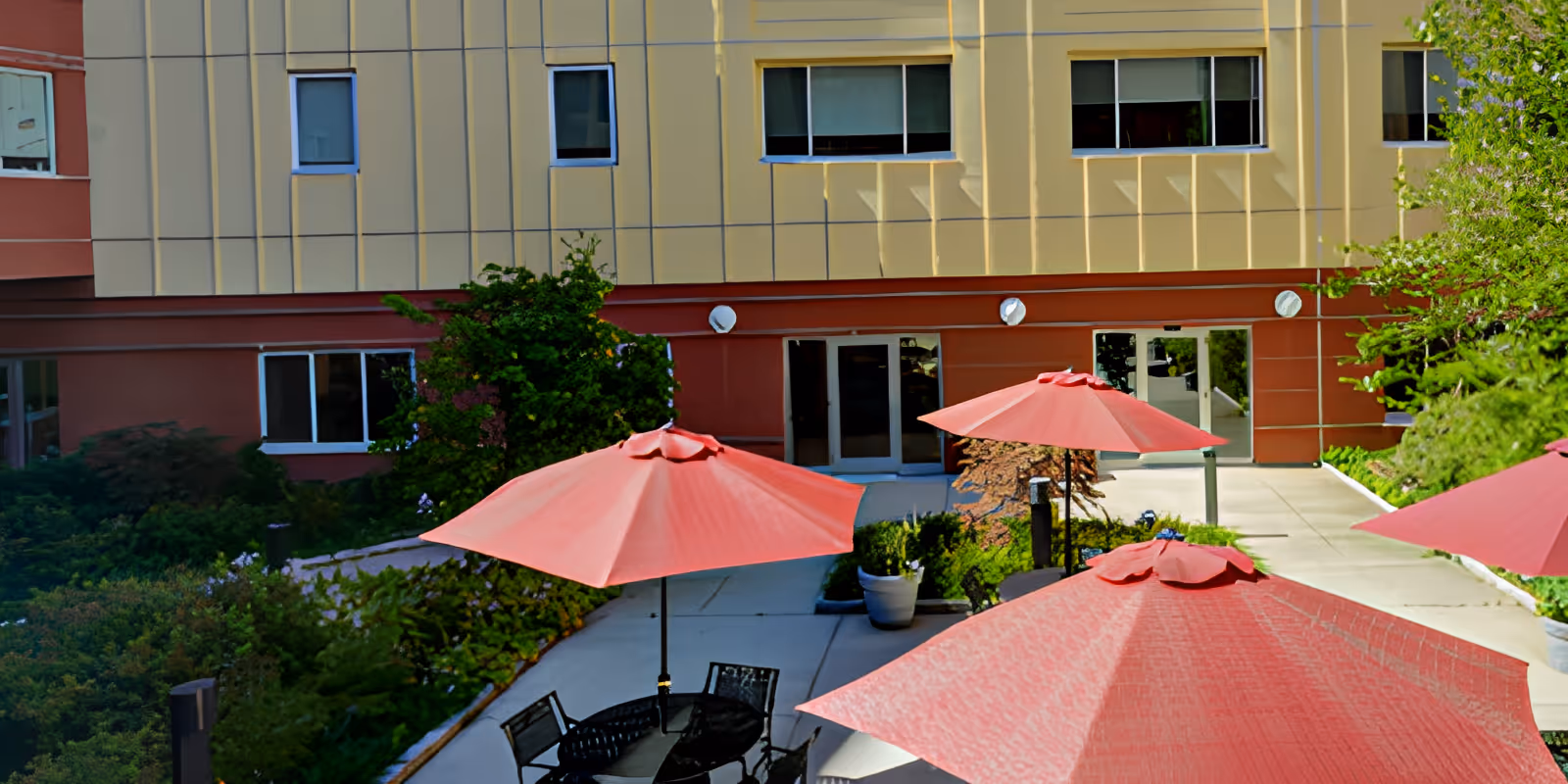 Courtyard with red patio umbrellas and round tables in front of a multi-story building with windows and glass doors.