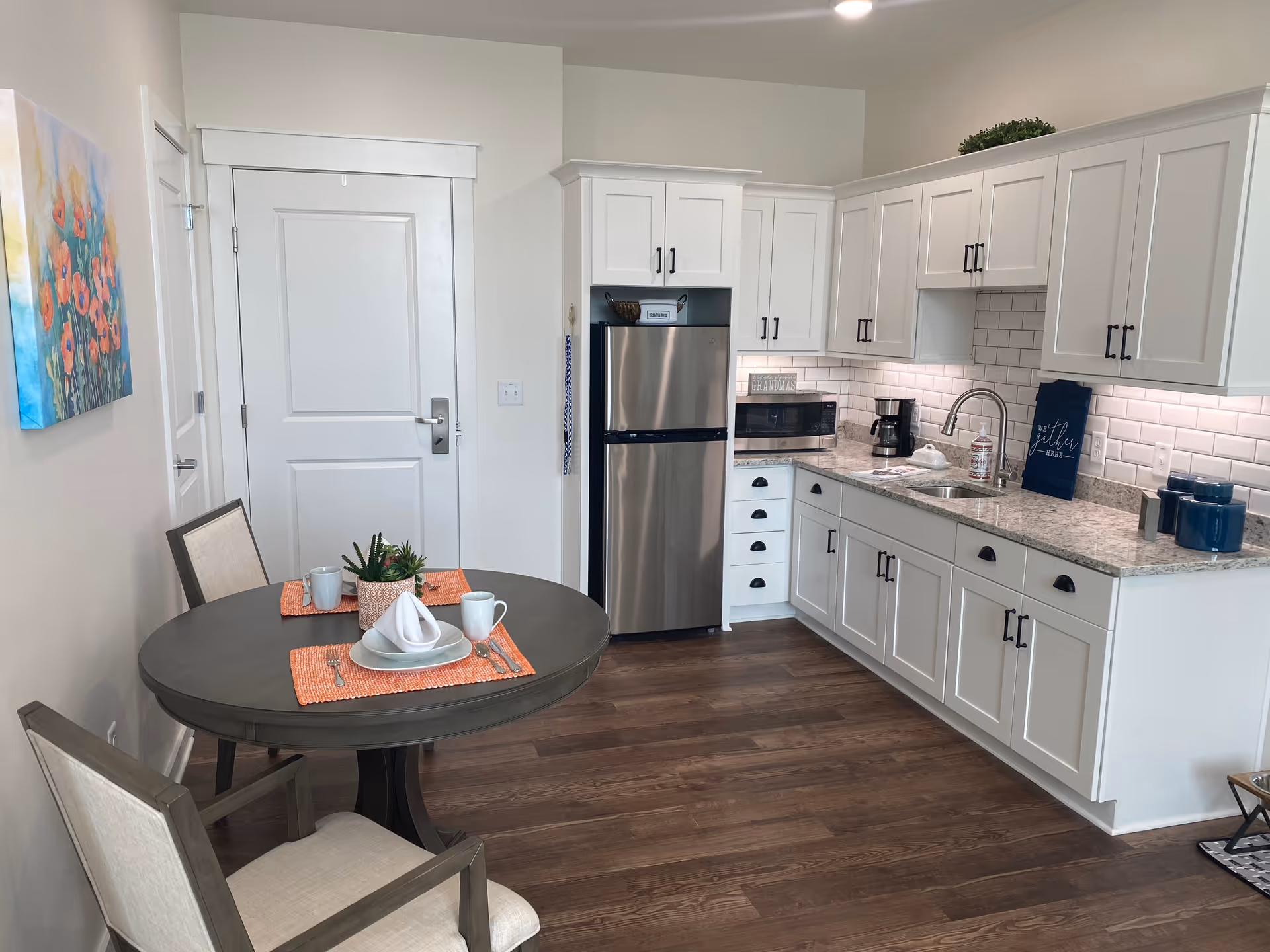 Bright kitchenette with white cabinets, stainless refrigerator, sink and a round dining table set with two chairs.