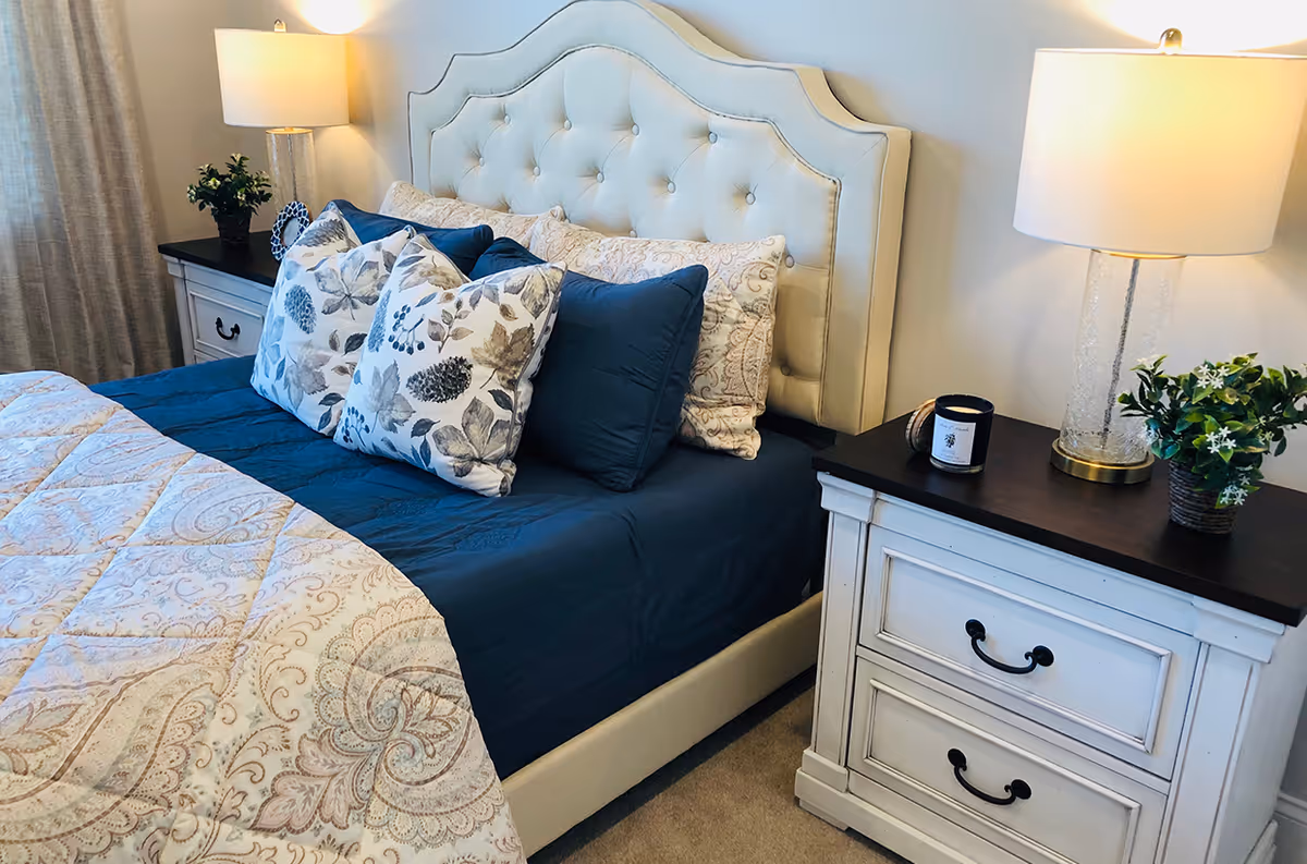 A neatly made bed with a tufted upholstered headboard, patterned pillows and navy bedding, flanked by a nightstand with a lamp and plant.