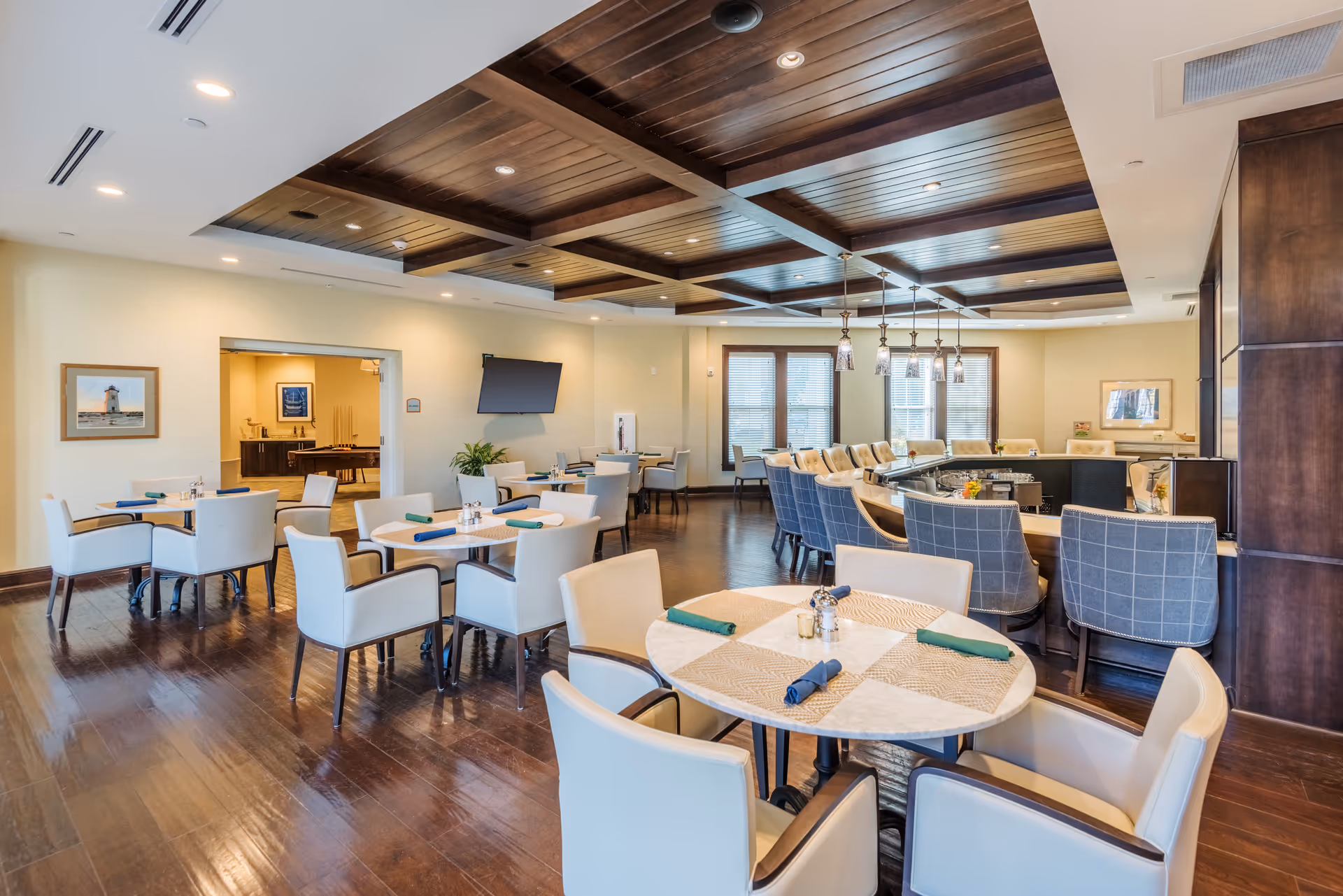 A spacious dining room with multiple tables and chairs arranged neatly. The room features a wooden coffered ceiling with recessed lighting, large windows letting in natural light, and a bar area with high-backed chairs. The walls are decorated with framed pictures, and there is a flat-screen TV mounted on one wall.