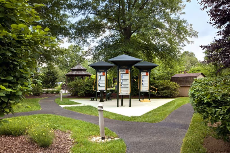 Landscaped outdoor courtyard with paved walking paths, three covered exercise stations, a gazebo, and surrounding trees and shrubs.