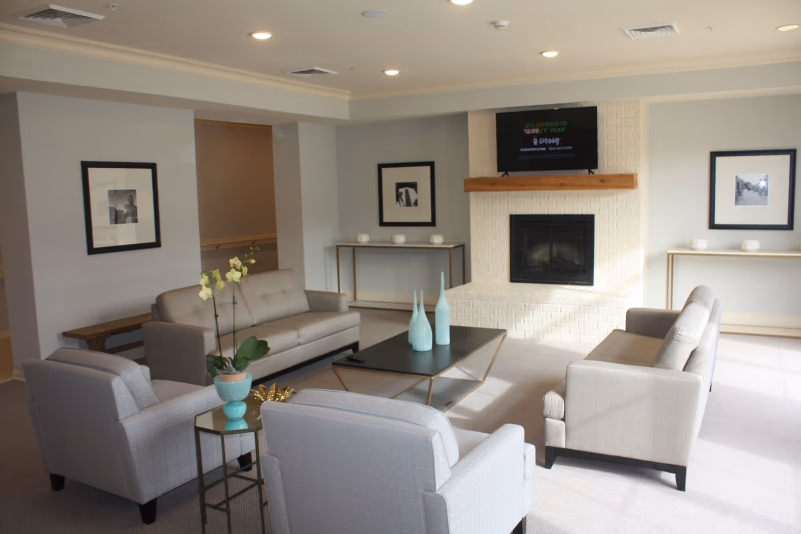 A modern living room with two gray armchairs and two gray sofas arranged around a black coffee table with three decorative vases. There is a white brick fireplace with a wooden mantel and a TV mounted above it. The room has light-colored walls, carpeted floor, framed black and white pictures on the walls, and two console tables with decorative items.