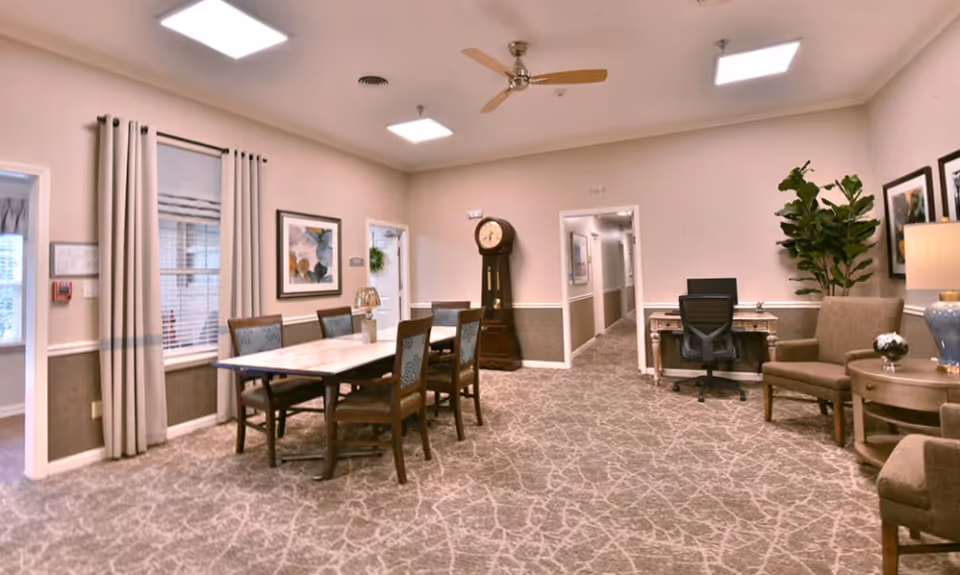 A well-lit interior room with a dining table and six chairs on the left side, a grandfather clock against the far wall, and a small desk with an office chair and a seating area with two armchairs and a side table with a lamp on the right side. The room has beige walls, patterned carpet, framed artwork, and a large potted plant near the seating area.