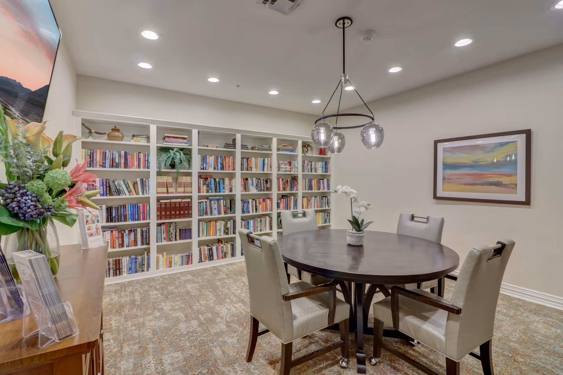 A cozy reading room with a round wooden table surrounded by four cushioned chairs. Behind the table is a large white bookshelf filled with books and decorative plants. The room is well-lit with recessed ceiling lights and a modern hanging light fixture. A colorful abstract painting hangs on the wall, and a wooden cabinet with a vase of flowers and brochures is visible on the left side.