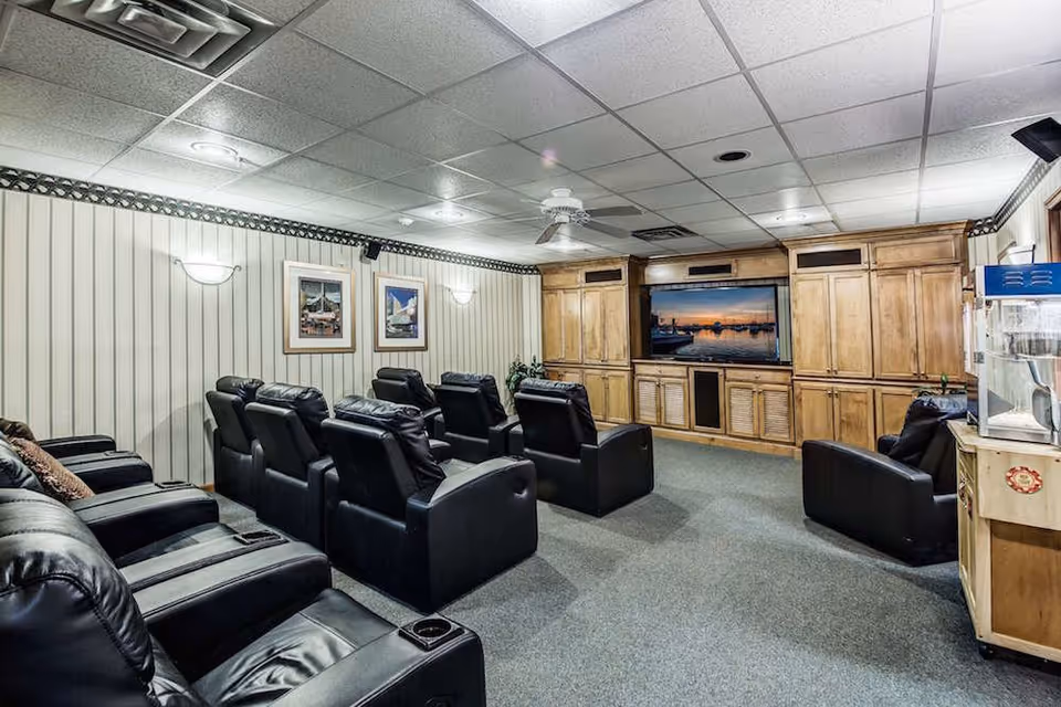 A cozy media room with multiple black leather recliners arranged in rows facing a large flat-screen TV mounted on a wooden entertainment center. The room has a carpeted floor, light-colored paneled walls with framed pictures, ceiling lights, and a ceiling fan. A popcorn machine is visible on the right side of the room.