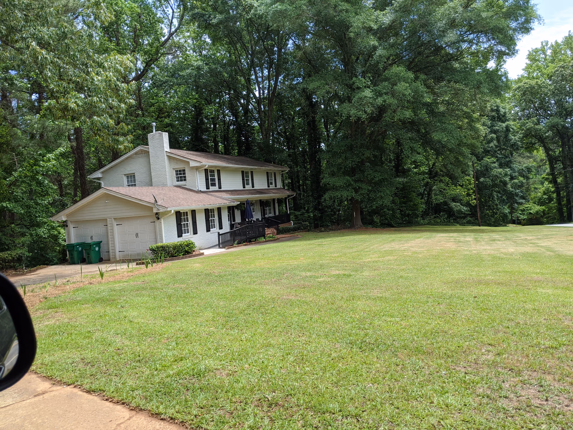 A two-story white house with a brown roof surrounded by large green trees and a well-maintained grassy lawn. The house has a front porch with a ramp and a double garage. There are two green trash bins near the garage. The sky is clear and blue.