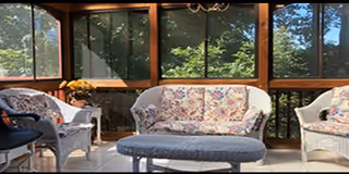 A sunlit enclosed porch or sunroom with large windows showing green trees outside. The room contains a floral-patterned cushioned sofa, two matching armchairs, and a cushioned ottoman in front of the sofa. There is a small table with a flower arrangement near one of the armchairs.