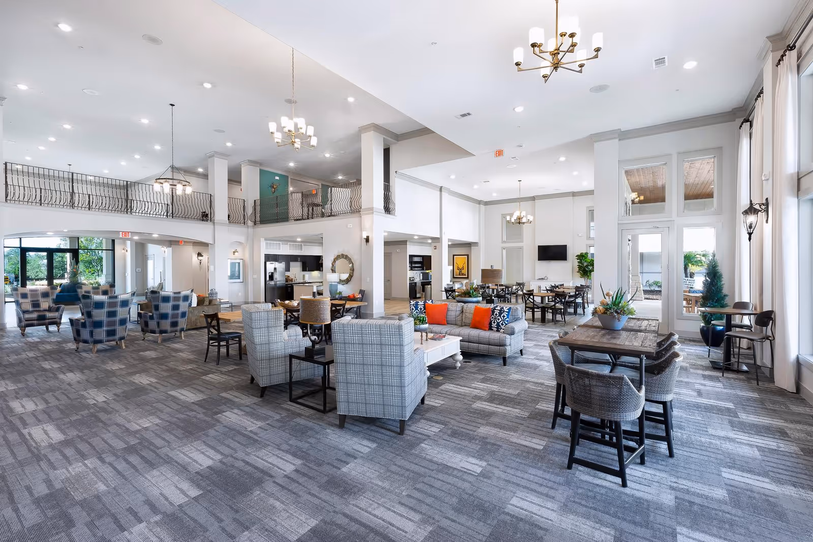 Spacious and well-lit senior living facility common area with modern furniture including sofas, armchairs, dining tables, and chairs. The room features high ceilings with chandeliers, large windows with curtains, and a balcony railing on the upper level. There is a kitchen area visible in the background and a TV mounted on the wall.