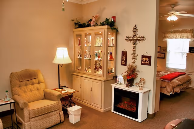 A cozy living area with a beige upholstered armchair next to a floor lamp and small side table. A glass-front cabinet displays decorative figurines. A white electric fireplace is positioned against the wall, adorned with a floral arrangement and small decorations. A large decorative cross hangs on the wall above the fireplace. In the background, a bedroom with a bed covered in beige bedding and a red blanket is visible through an open doorway.