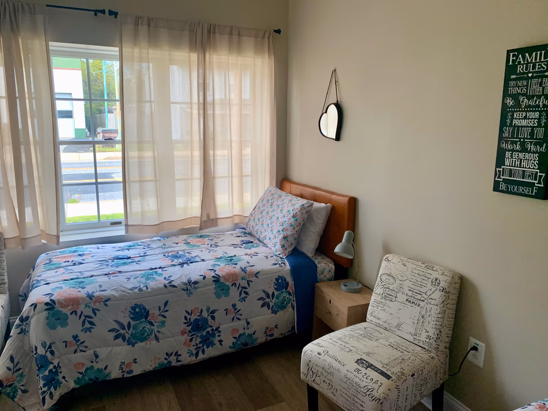 A small bedroom with a single bed covered in floral bedding, a wooden nightstand with a small lamp, a cushioned chair with script print, a heart-shaped mirror hanging on the wall, and a window with sheer curtains letting in natural light.