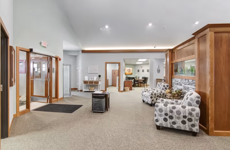 Spacious senior living facility lobby with patterned armchairs, wooden paneling, entrance doors, and a seating area leading to interior rooms.