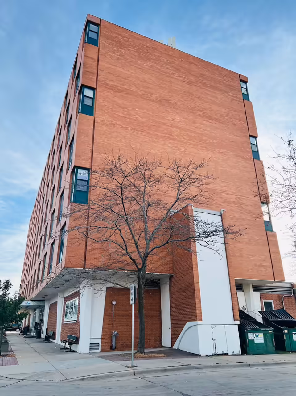 A tall brick building with multiple windows, a leafless tree in front, and a sidewalk with benches and a street sign. There are green dumpsters on the right side near the building.